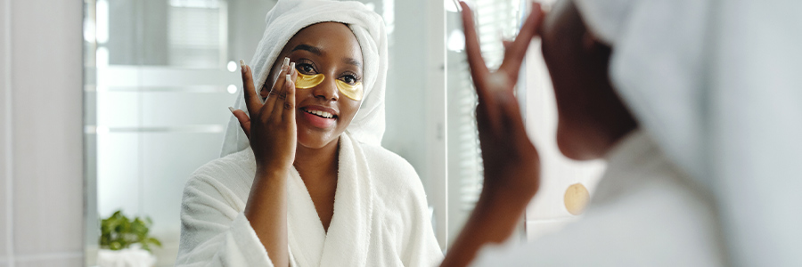Une personne portant un peignoir de bain regarde dans un miroir de salle de bain en appliquant un produit de soins pour le visage.