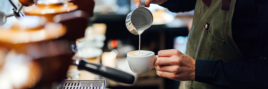 Derrière le comptoir d’un café, un barista verse de la mousse de lait dans une tasse pour préparer un café au lait.