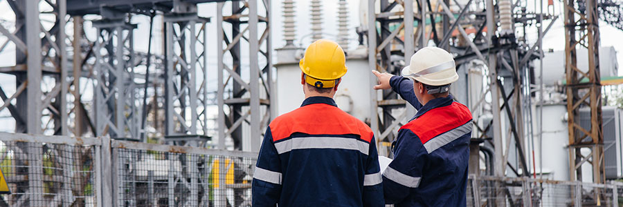 Deux travailleurs portant un casque de sécurité dans une station de transfert d’électricité, entourés de transformateurs à haute puissance.