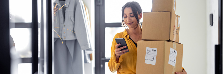 Une femme transporte plusieurs boîtes de livraison à sa porte avant et regarde son téléphone cellulaire.