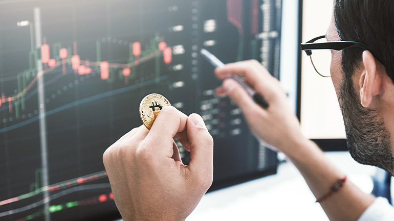 Un investisseur tient un jeton physique de bitcoins tout en regardant des graphiques financiers sur ses écrans d’ordinateur.